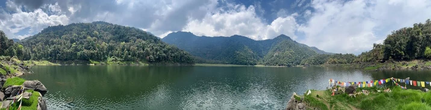 A panorama of Khabji Hoka Tso, still water ringed by forested hills, prayer flags along the near shore