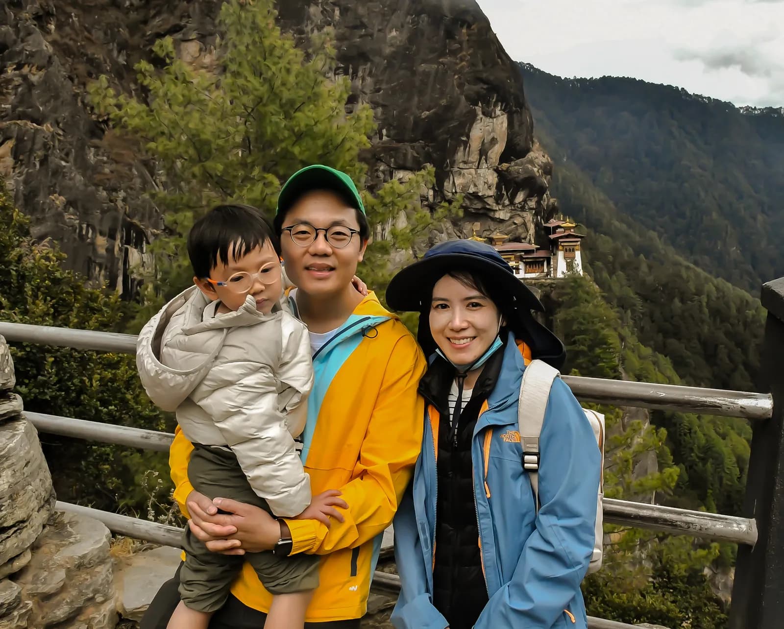 A family at the railing on the Taktsang trail, the monastery small on the rock face beyond