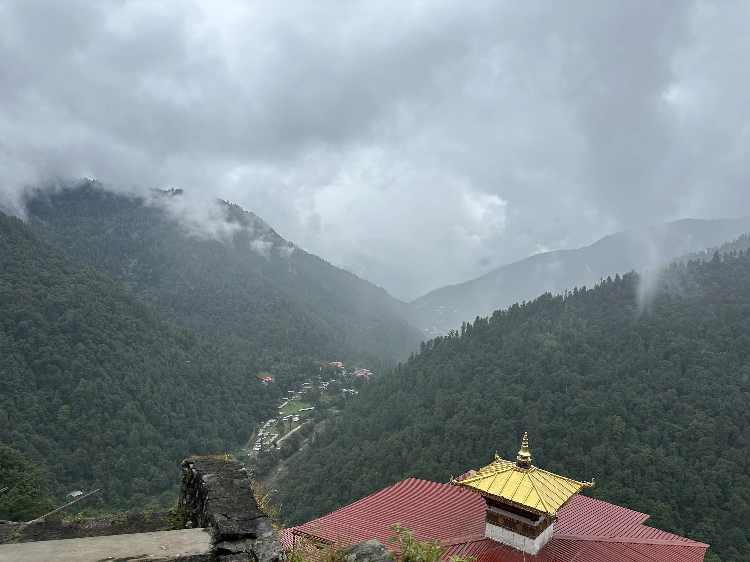 A forested Himalayan valley under low cloud, with a gilded temple roof in the foreground