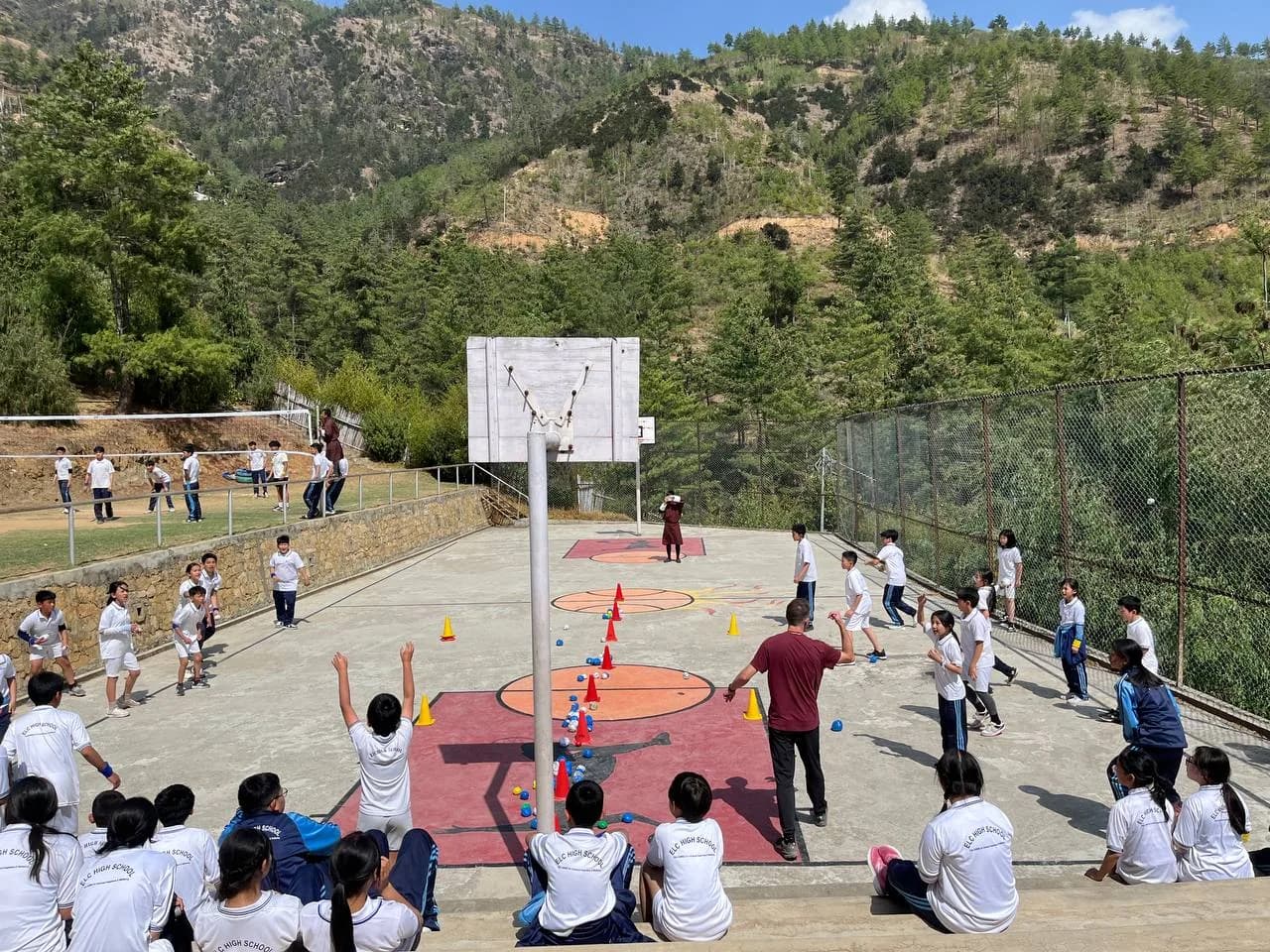 ELC High School students playing basketball on the school's court, with a coach and the Thimphu hills behind